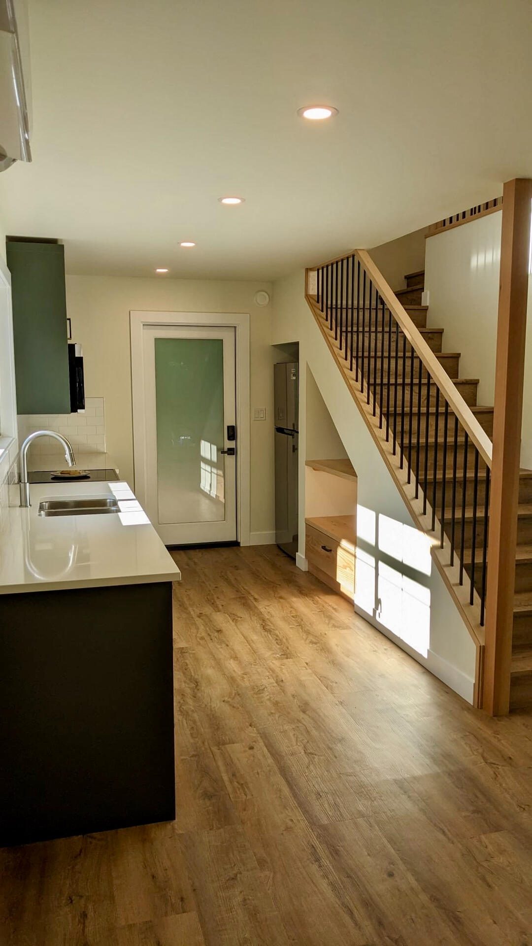 Open-concept interior showing kitchen counter, wood stairwell with built-in under-stair storage nook, and modern front door.
