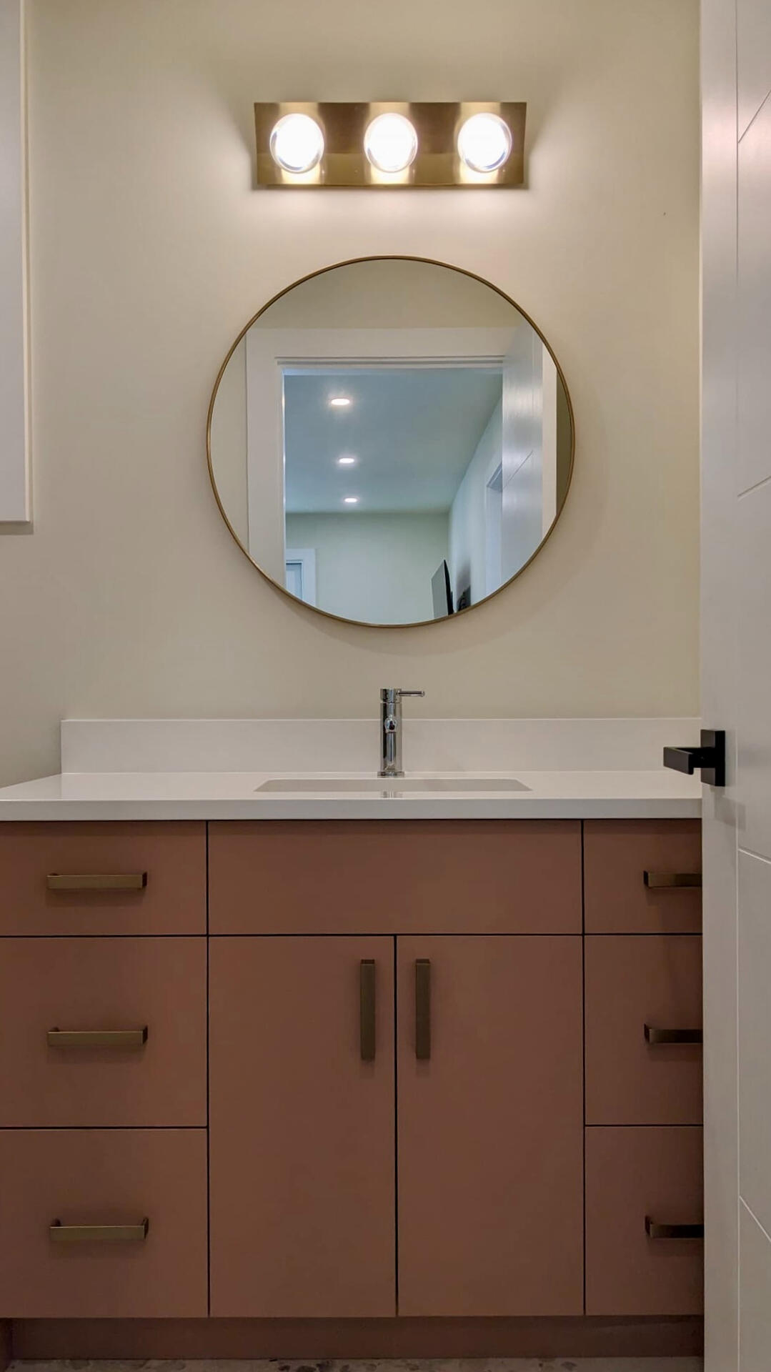 Terracotta bathroom vanity featuring a white quartz countertop, circular gold frame mirror, and modern three-light vanity bar.