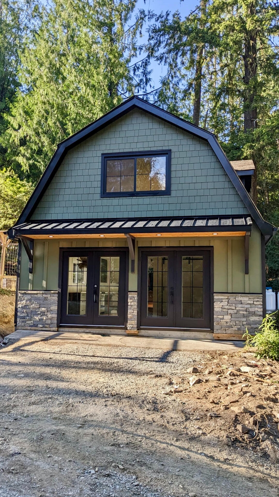 Exterior view of a two-story modern carriage home with green vertical siding, stone veneer accents, and glass double doors surrounded by trees.